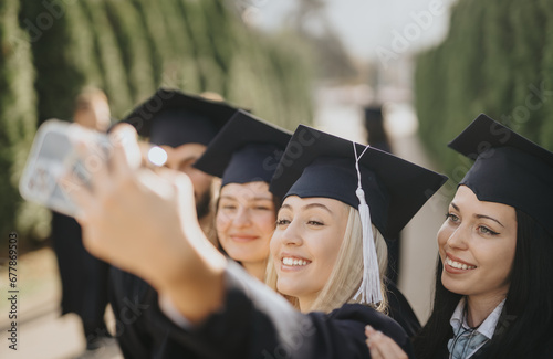 University students celebrate graduation in a sunny park. Smiling, they take selfies, and share happiness. Achievements and success create positive memories.