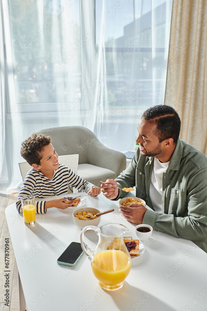vertical shot of joyful african american father and son enjoying breakfast and smiling happily vertical shot of joyful african american father and son enjoying breakfast and smiling happily