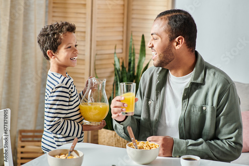 cheerful african american boy with jug of orange juice smiling at his father at breakfast table