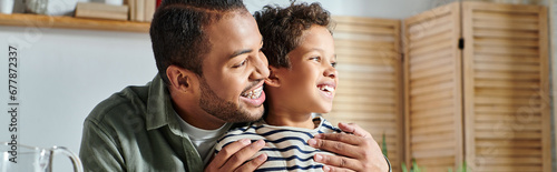 cheerful african american father and son in casual homewear hugging and looking away, banner