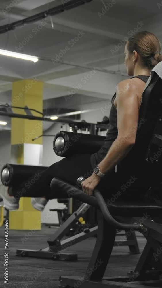 Vertical video. Young Woman in Front of a Mirror Doing Leg Extensions on a Machine. Sports Training and Workout Program for Women. Vertical video. Young Woman in Front of a Mirror Doing Leg Extensions on a Machine. Sports Training and Workout Program for Women.