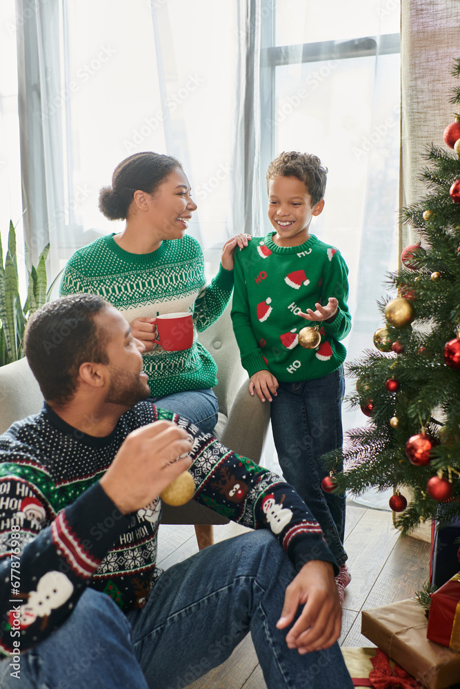 african american woman with coffee cup looking at her husband and son decorating Christmas tree african american woman with coffee cup looking at her husband and son decorating Christmas tree