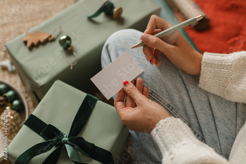 Close-up of a woman sitting cross-legged on the floor next to a wrapped Christmas gift writing a Christmas card photo