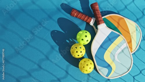 image from above of two pickleball paddles on the floor of a blue court on which three pickleball balls appear rolling
