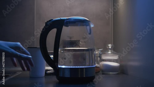 beautiful young female hand turns on a transparent glass electric kettle with water presses the button blue lamp lights up at dusk photo