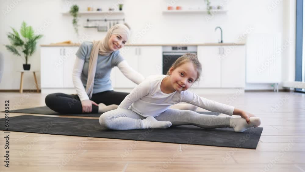 Slim muslim lady on yoga mat stretching arms and legs while training with small female kid. Young parent and daughter in sportswear spend time happily during workout. Slim muslim lady on yoga mat stretching arms and legs while training with small female kid. Young parent and daughter in sportswear spend time happily during workout.