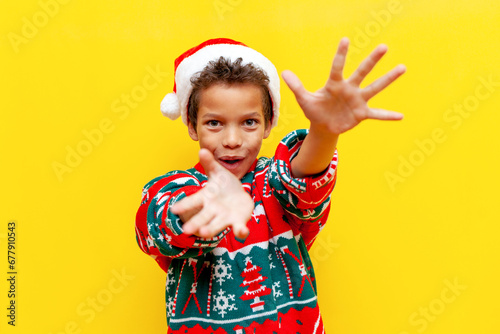 african american boy in christmas sweater and santa hat gives and throws on yellow isolated background