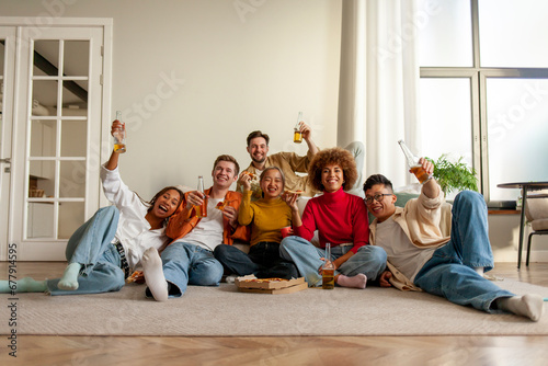 multiracial group of young friends sitting at home on the sofa with bottles of beer and eating pizza and watching TV