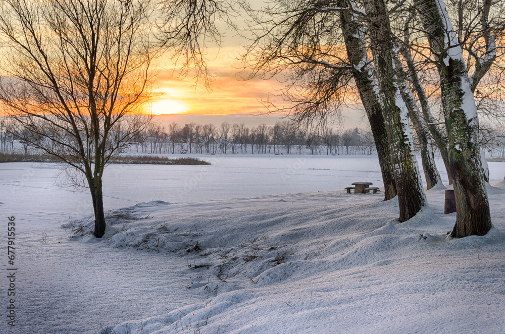 Snowy winter landscape with sunset in the background. The frozen Tisza backwater can be seen in Tiszalök, Hungary. Bare trees and branches are still visible in the picture. Hungarian rural landscape. 