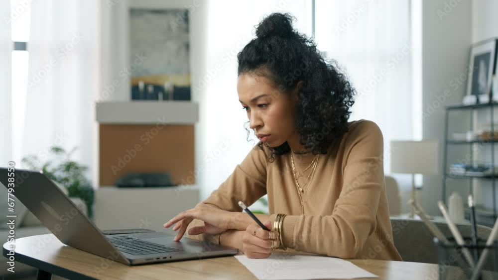 Young multi racial woman working remotely at home office looks at laptop computer, e learning online on internet website. Concentrated focused african female student makes notes in work document 4K Young multi racial woman working remotely at home office looks at laptop computer, e learning online on internet website. Concentrated focused african female student makes notes in work document 4K
