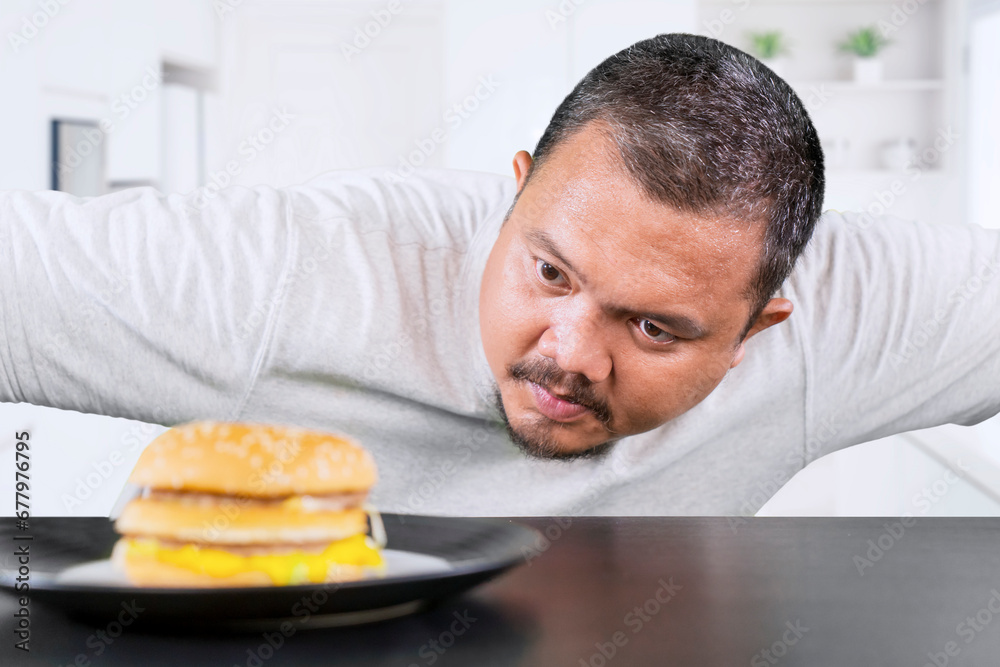 Middle aged overweight man looking at a cheeseburger thinking of eating it at home Middle aged overweight man looking at a cheeseburger thinking of eating it at home