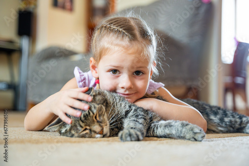 Portrait of cute little girl with gray cat petting pet and lying on carpet in home living room