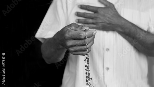Praying rosary concept with man holding rosary beads in hand and another hands on chest in black and white background. Catholic religious symbol of faith. Devotion prayer to Mother Mary.