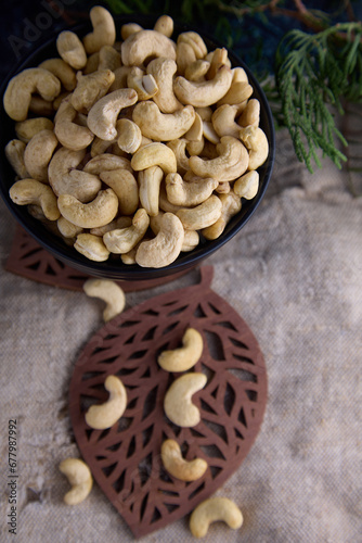 Plate of cashews stands on a figured wooden sheet on burlap. Black plate filled with cashew nuts against the background of old burlap and a coniferous green branch.