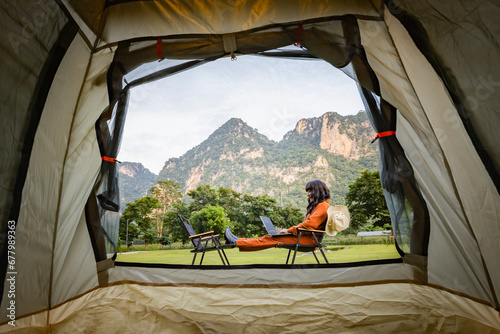Young woman sitting near tent with bucket hat hanging on camping chair, using laptop and working remotely while camping in nature during adventure trip in forest. View of hills covered by forest. 
