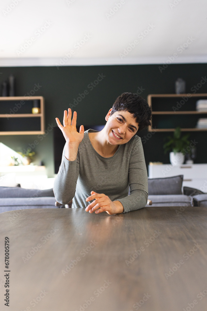 Fototapeta premium Happy biracial woman sitting at dining table having video call at home, waving, copy space