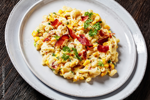 mac and cheese with corn, bacon topped with panko breadcrumbs on plates with fork on dark wooden table, dutch angle view, close-up