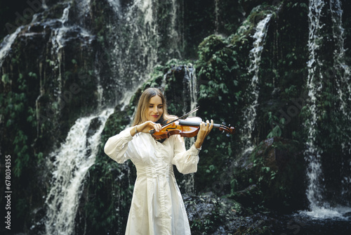 Beautiful Caucasian woman playing violin near waterfall. Music and art concept. Female wearing white dress in nature. Water splash. Tropical forest. Banyu Wana Amertha waterfall, Bali. photo