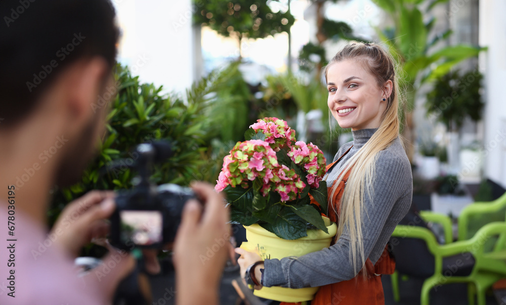 Happy Vlogger Posing with Flower to Camera Man. Beautiful Blonde Girl Holding Colorful Blooming Hydrangea in Flowerpot. Smiling Woman Florist Recording Hortensia Bloom for Domestic Plant Vlog