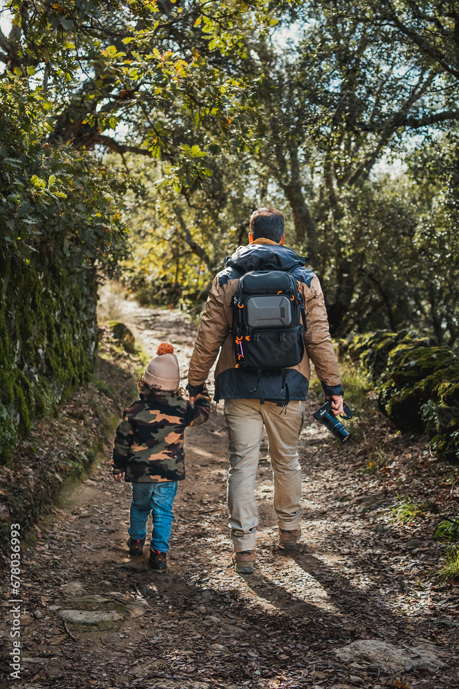 Photographer father with backpack and fashionable son, walking along a beautiful forest path full of dry leaves and surrounded by green, vertical