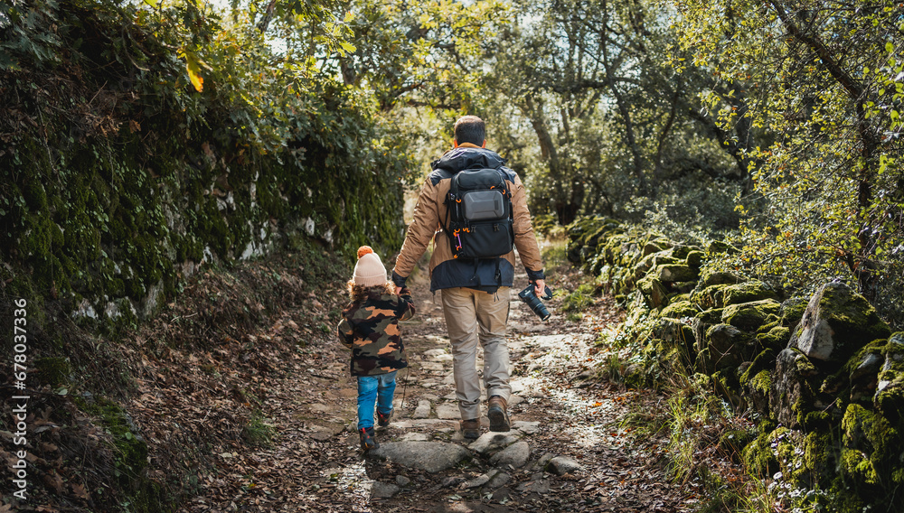Photographer father with backpack and fashionable son, walking along a beautiful forest path full of dry leaves and surrounded by green Photographer father with backpack and fashionable son, walking along a beautiful forest path full of dry leaves and surrounded by green