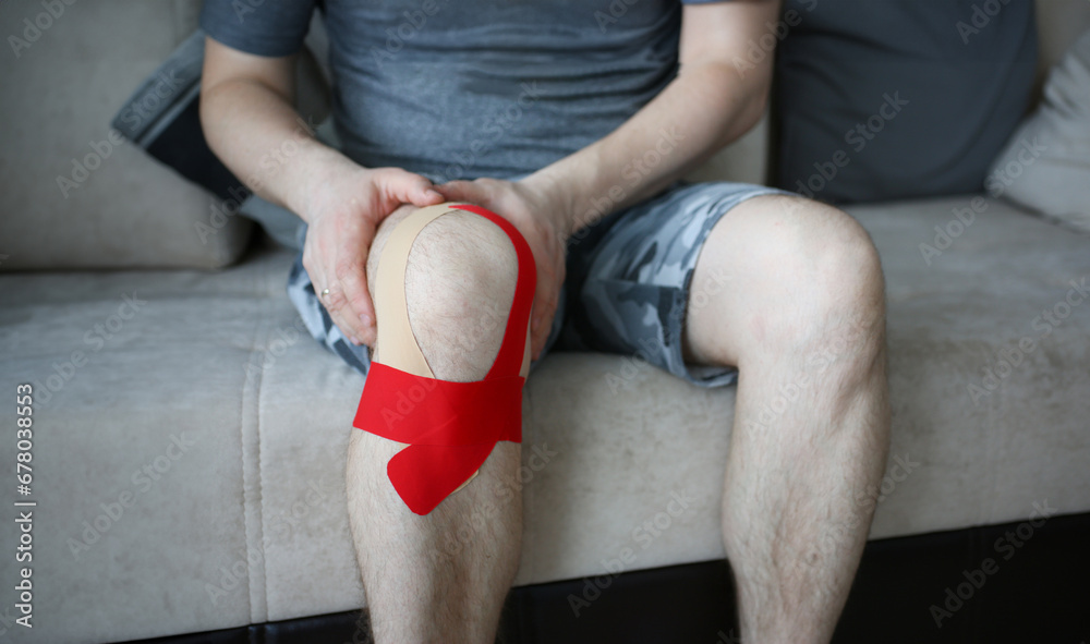 Naklejka premium Troubled male holding tied up knee in pain. Man sitting on soft sofa and suffering from unbearable discomfort. Medication treatment and healthcare concept