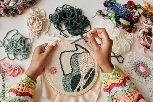 Above view of hands of young female designer making creative embroidery on piece of white canvas clamped in hoop