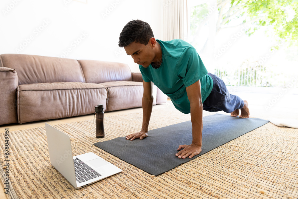 Biracial man exercising on mat and using laptop at home