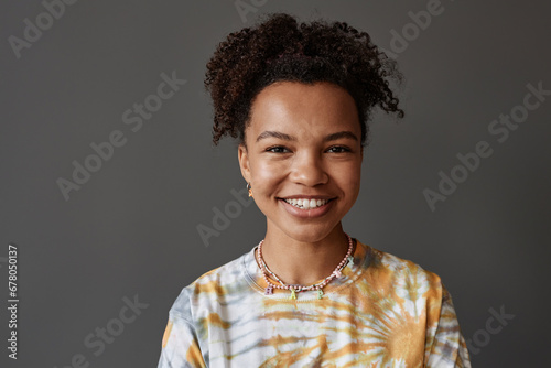Young smiling African American woman in tie dye t-shirt looking at camera while standing in isolation over grey background in studio