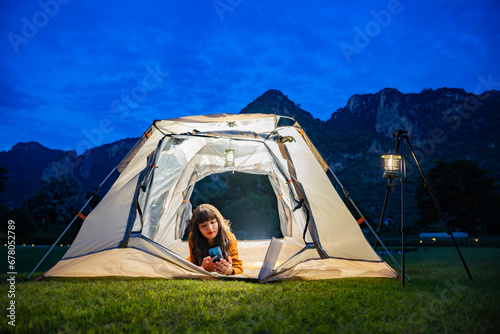 Charming asian woman with beautiful smile reading good news on mobile phone while lying in tent and warm night light under dark blue sky twilight time. The tent lights at night from the lamp camping.