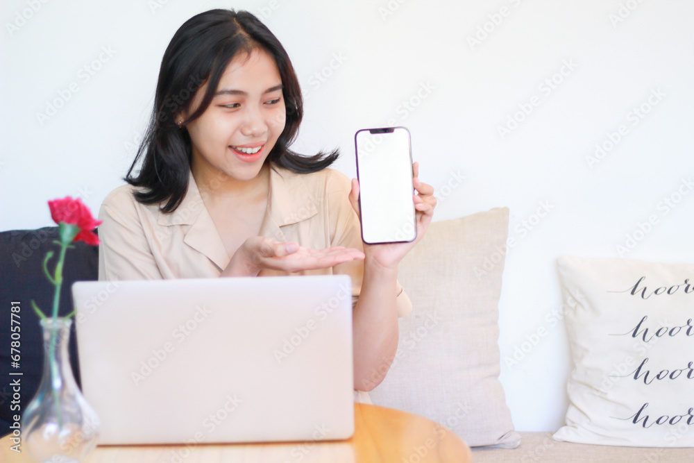 excited asian woman sitting in the cozy couch wearing casual clothes showing white screen of smartphone for product application display excited asian woman sitting in the cozy couch wearing casual clothes showing white screen of smartphone for product application display