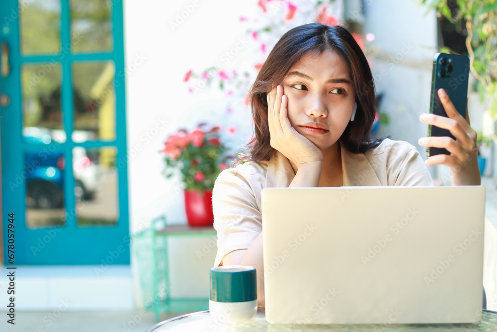 confused asian woman holding smartphone with laptop on table sitting in outdoors cafe working space wearing casual clothes - obrazy, fototapety, plakaty 