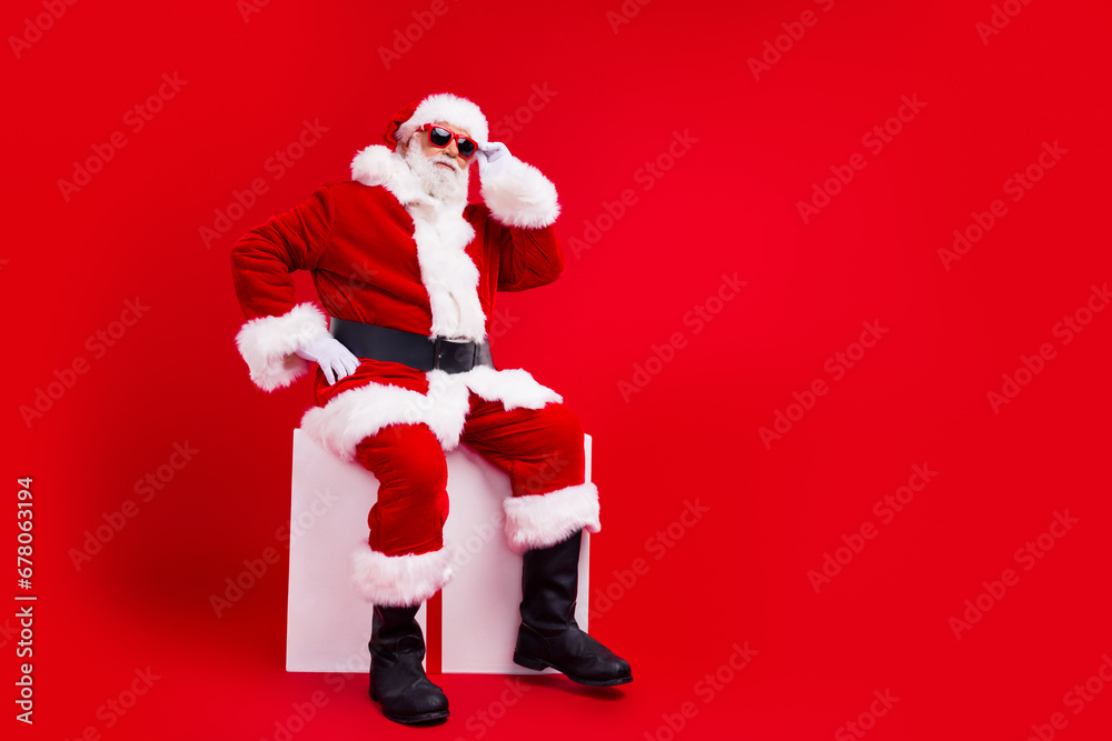 Full length photo of cheerful retired man santa claus sitting cube christmas event offer empty space isolated on red color background Full length photo of cheerful retired man santa claus sitting cube christmas event offer empty space isolated on red color background