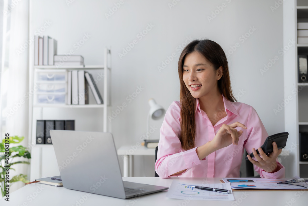 Young Asian woman sitting at desk with data and graph paper using laptop, analyzing accounts, planning strategies on business improvement in office. Young Asian woman sitting at desk with data and graph paper using laptop, analyzing accounts, planning strategies on business improvement in office.