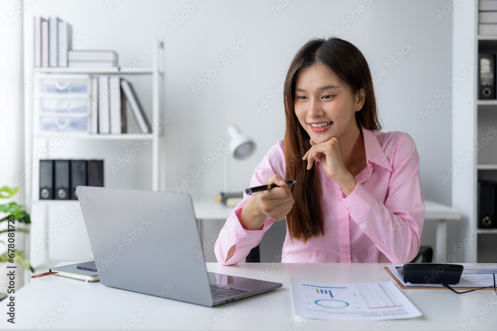 Young Asian woman sitting at desk with data and graph paper using laptop, analyzing accounts, planning strategies on business improvement in office. Young Asian woman sitting at desk with data and graph paper using laptop, analyzing accounts, planning strategies on business improvement in office.
