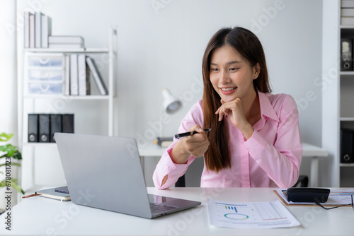 Young Asian woman sitting at desk with data and graph paper using laptop, analyzing accounts, planning strategies on business improvement in office.