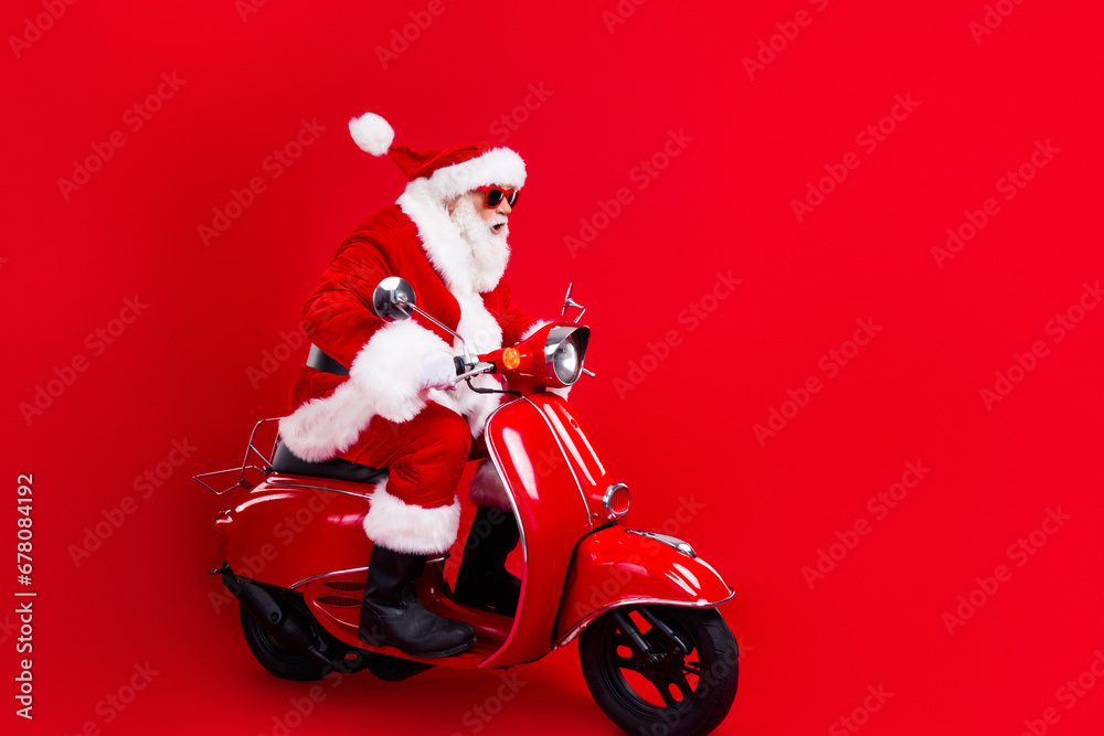 Full length photo of excited santa claus with white beard in sunglass driving scooter look empty space isolated on red color background Full length photo of excited santa claus with white beard in sunglass driving scooter look empty space isolated on red color background