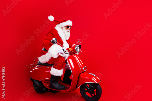 Full length photo of excited santa claus with white beard in sunglass driving scooter look empty space isolated on red color background