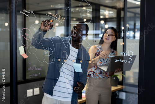Happy diverse male and female colleague brainstorming, writing on glass wall, using laptop in office photo