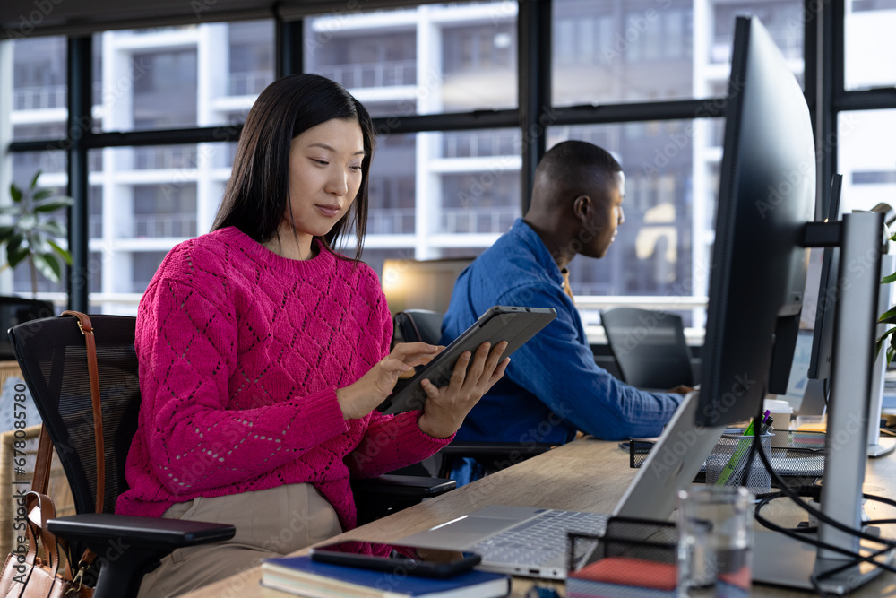 Focused asian casual businesswoman using tablet at desk with diverse colleague in office