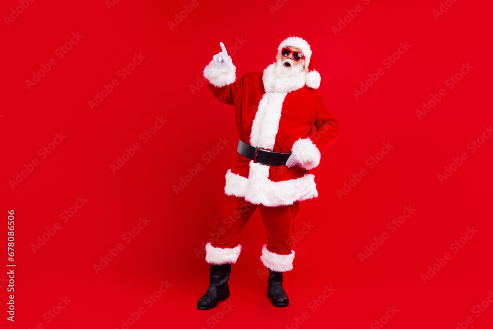 Portrait of ecstatic funny santa claus with white beard in sunglass directing at billboard empty space isolated on red color background