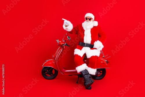 Full size photo of positive grandfather wear santa costume sit on moped indicating at promo empty space isolated on red color background