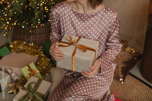 Stylish young woman in a silk dress sitting on the floor next to a Christmas tree holding a wrapped Christmas gift photo