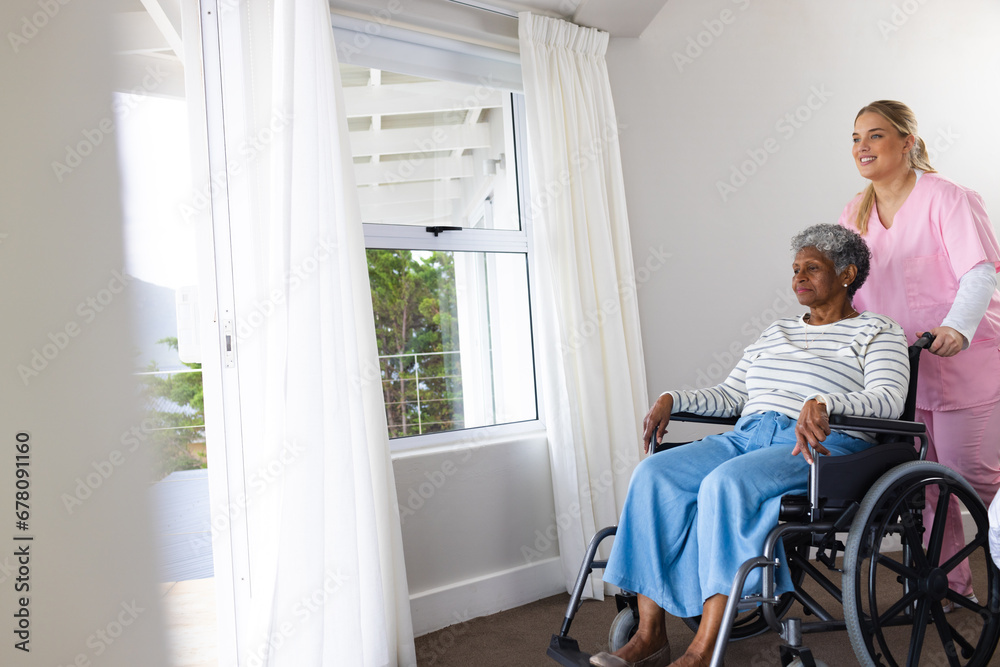 Caucasian female nurse and senior african american female patient in wheelchair by window Caucasian female nurse and senior african american female patient in wheelchair by window