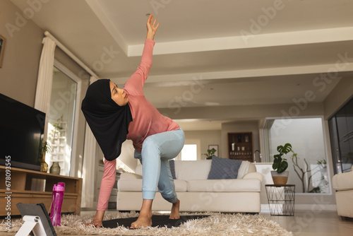 Biracial woman in hijab practicing yoga on mat using tablet at home photo