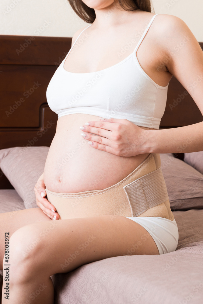 closeup pregnant woman dresses bandage on belly at home on bed. Orthopedic abdominal support belt closeup pregnant woman dresses bandage on belly at home on bed. Orthopedic abdominal support belt