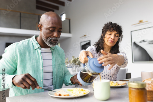 Happy mature diverse couple having breakfast and pouring coffee in sunny kitchen