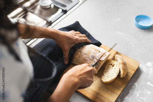 High angle of mature caucasian woman cutting fresh bread on breadboard in sunny kitchen, copy space photo