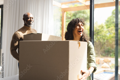 Happy diverse mature couple carrying packing boxes into sunny new home photo