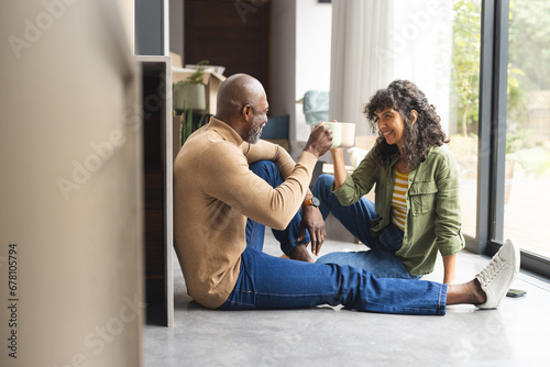 Happy diverse mature couple on floor toasting with coffees in sunny new home, copy space photo